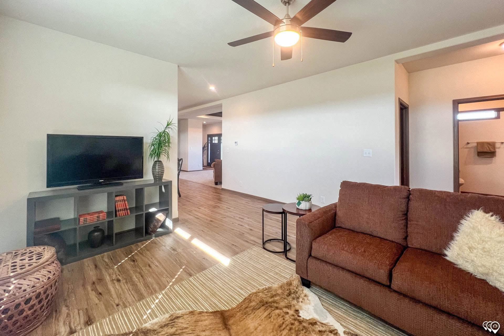 Modern living room with brown couch, beige walls, and wooden floors. A ceiling fan hangs above, and a TV sits on a shelf unit with decor items. Warm and inviting atmosphere.