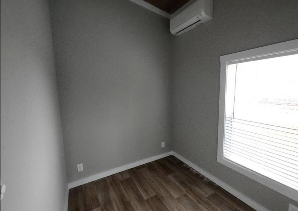 A small, empty room with gray walls, wooden floor, and a large window with blinds. An air conditioner is mounted near the ceiling, creating a minimalist feel.