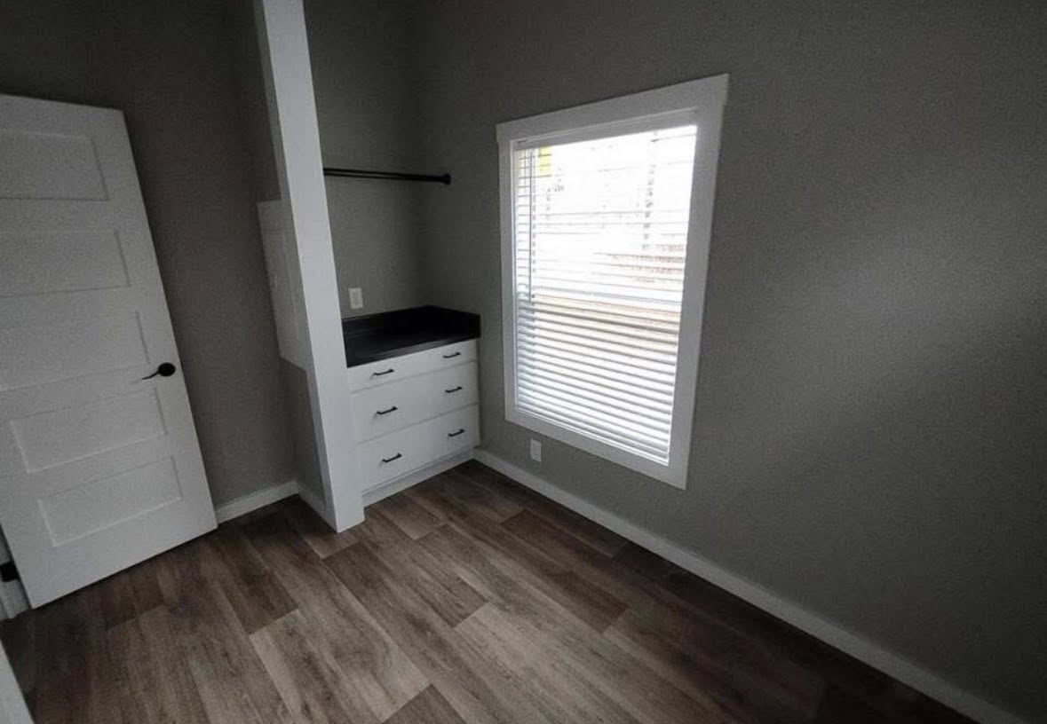 Small, grey-walled room with wood flooring, a window with blinds, and a narrow closet with white drawers. An open white door is visible.