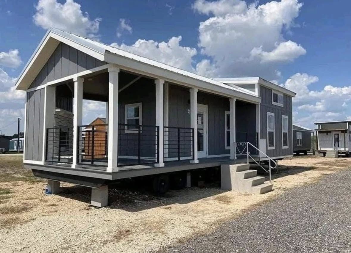 Small grey cabin with white trim and a porch, set on a sunny lot with a few other similar cabins in the background. Fluffy clouds fill the blue sky.