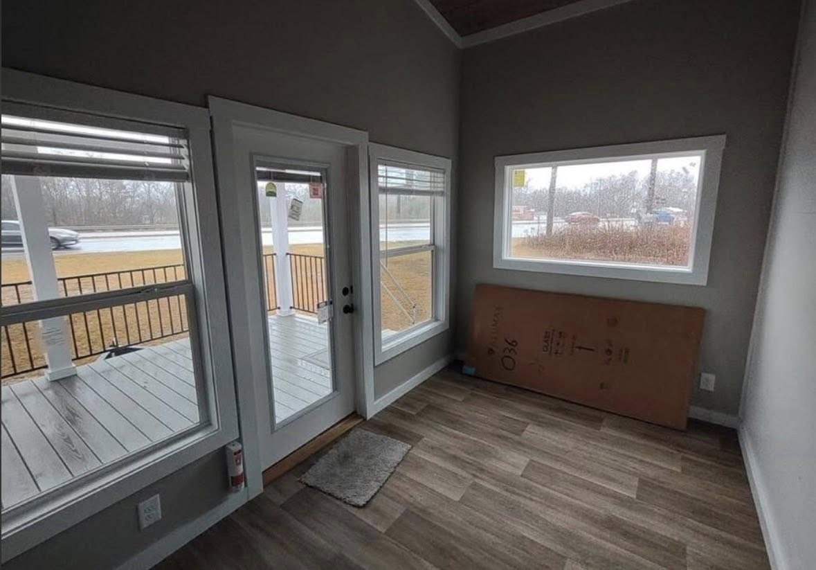 Bright room with wood flooring, large windows, and glass door leading to a porch overlooking a road and grassy area. A panel leans against the wall.