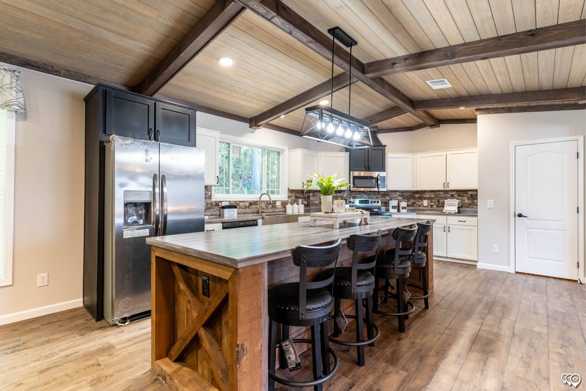 Spacious kitchen with rustic wooden beams, large island, and black bar stools. Stainless steel appliances and bright lighting create a cozy atmosphere.