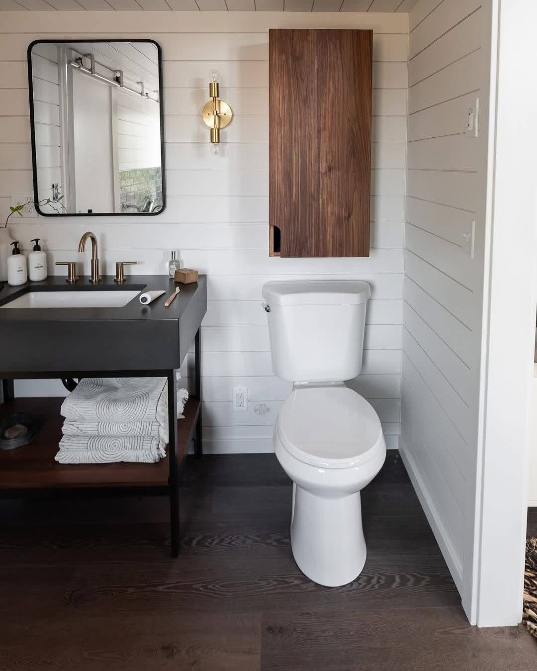 A modern bathroom features a white toilet, dark vanity with a rectangular mirror, brass faucet, and folded towels. Wood paneling and bright lighting create a clean, contemporary feel.