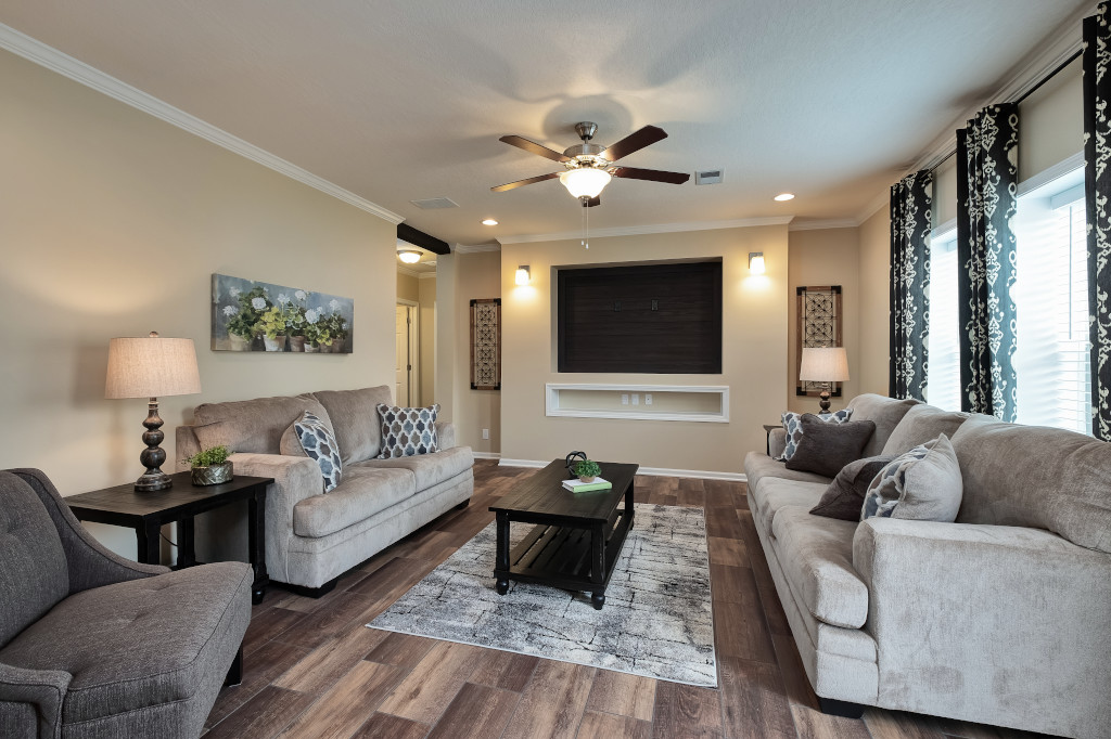 Warm living room with beige walls, wood flooring, and a ceiling fan. Features two sofas, a coffee table, and curtain-lined windows, creating a cozy, inviting atmosphere.