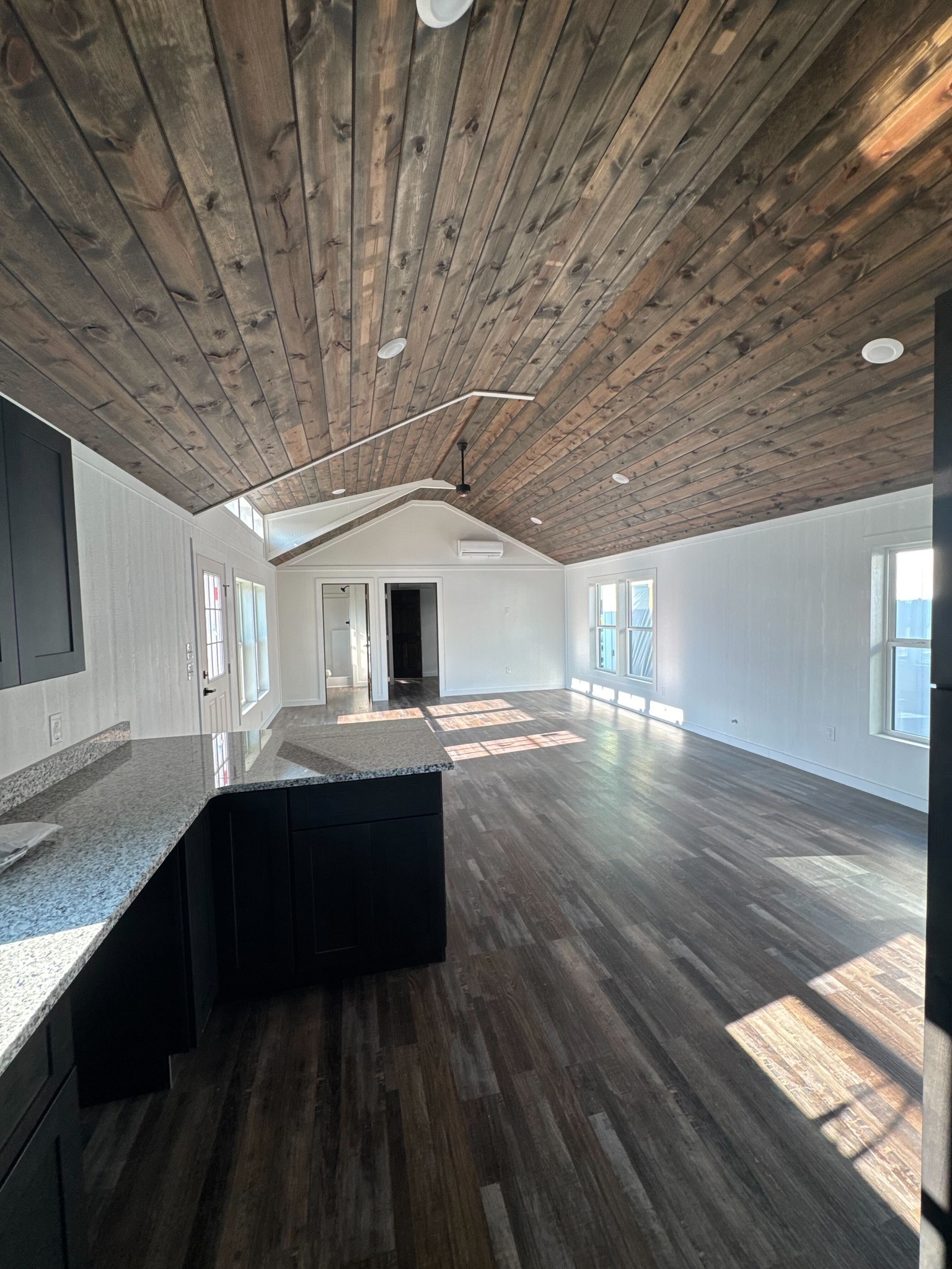 Spacious open-plan room with dark wood ceiling, matching floor, and white walls. Kitchen with granite countertops and natural light from large windows.
