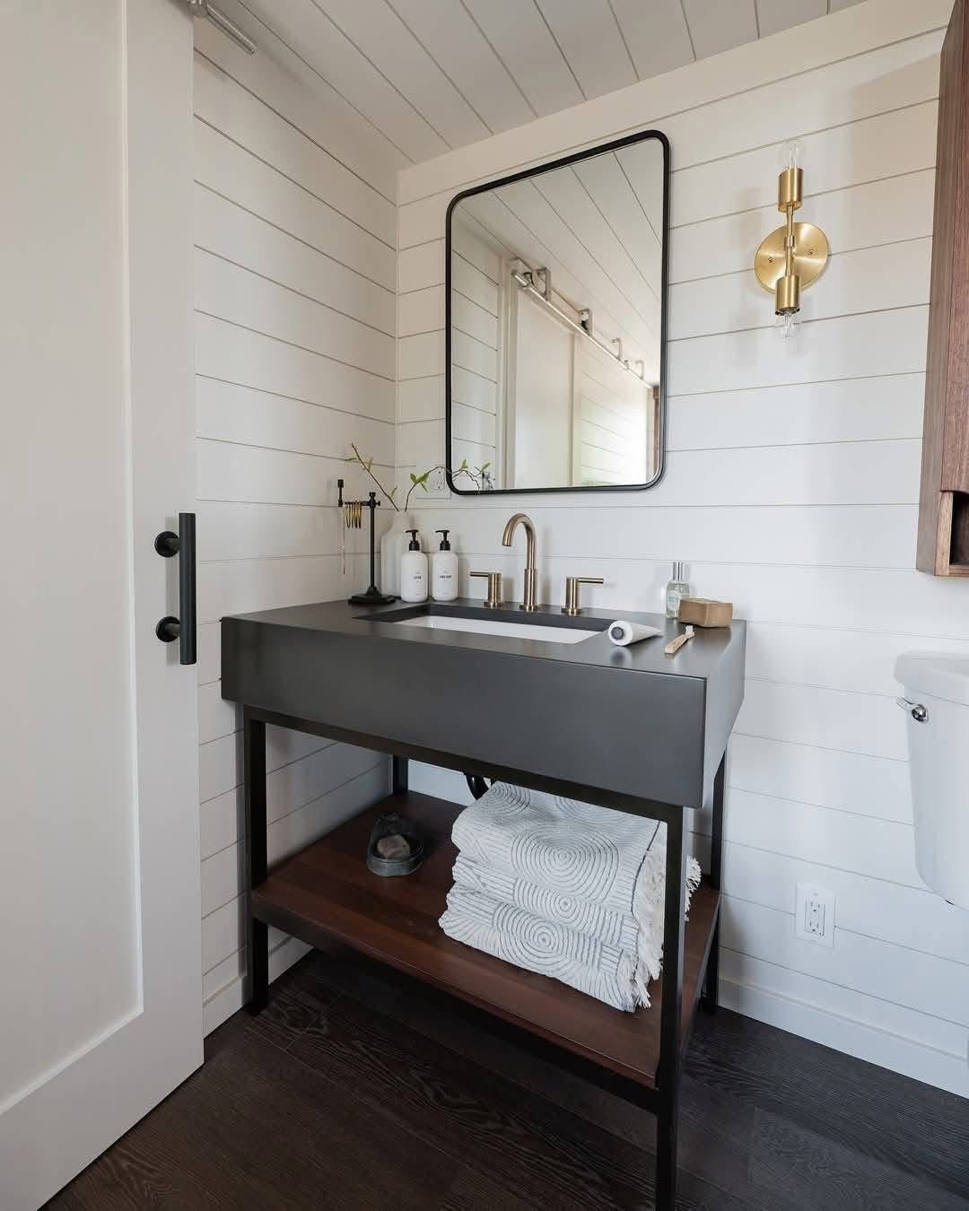 Stylish bathroom with a sleek black sink, gold fixtures, and a circular sconce. White shiplap walls and neatly folded towels create a modern, clean look.