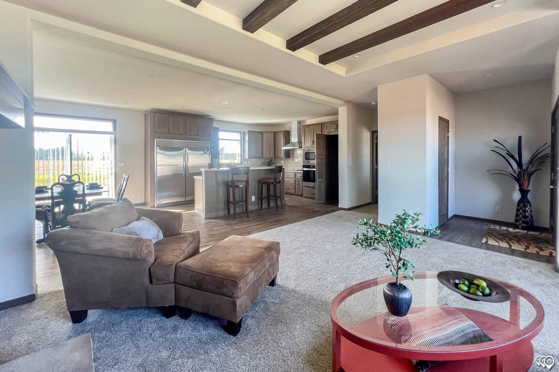 Spacious open-concept living area with beige walls and wood ceiling beams. Features a brown armchair, red coffee table, and modern kitchen and dining space.