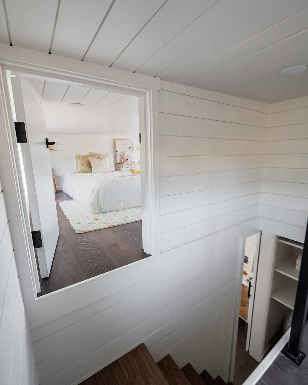 View from the top of a staircase in a cozy, white-paneled home. An inviting bedroom with a bed, pillows, and a rug is visible through an open door.