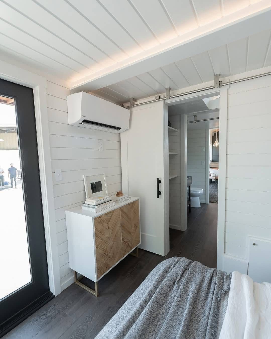Bright, modern tiny home interior with white shiplap walls, a sliding barn door, and minimalist furniture. Air conditioner and soft lighting create a cozy tone.