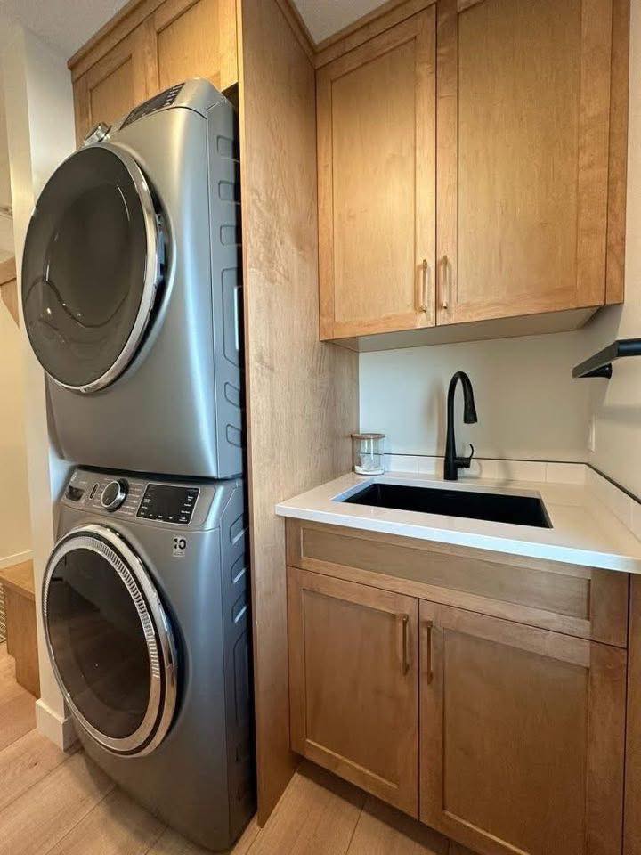 Stacked silver washer and dryer next to a wooden cabinet and countertop. A black faucet with a glass jar adds contrast, creating a clean, modern laundry area.