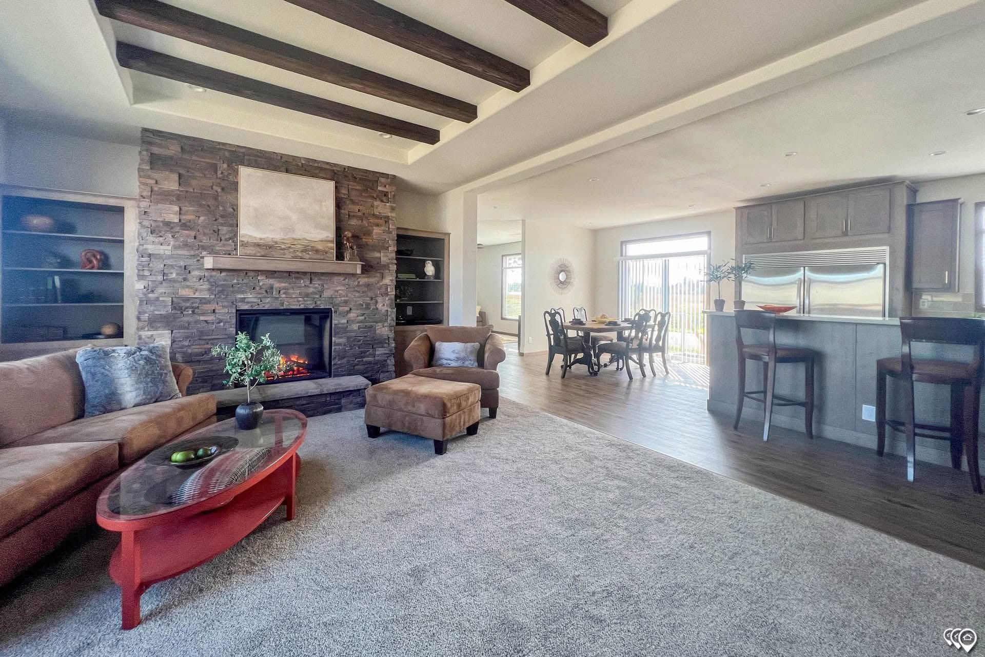 Spacious living room with a stone fireplace, wooden ceiling beams, and cozy brown furniture. A red coffee table adds vibrancy. Open kitchen and dining area.