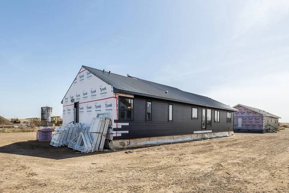 Partially constructed house with dark siding and a roof, surrounded by bare earth. Construction materials and equipment are visible, suggesting an in-progress build.