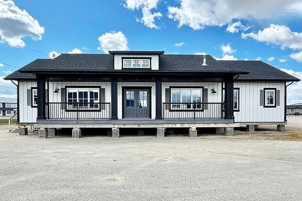 A modern, elevated white house with black trim and doors sits under a blue sky with clouds. The structure is supported by concrete blocks, creating a clean, contemporary look.