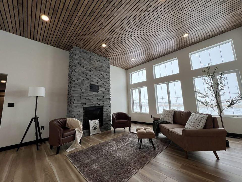 Modern living room with tall windows and wooden ceiling. Features a gray stone fireplace, brown leather chairs, a sofa, and a patterned rug. Cozy ambiance.