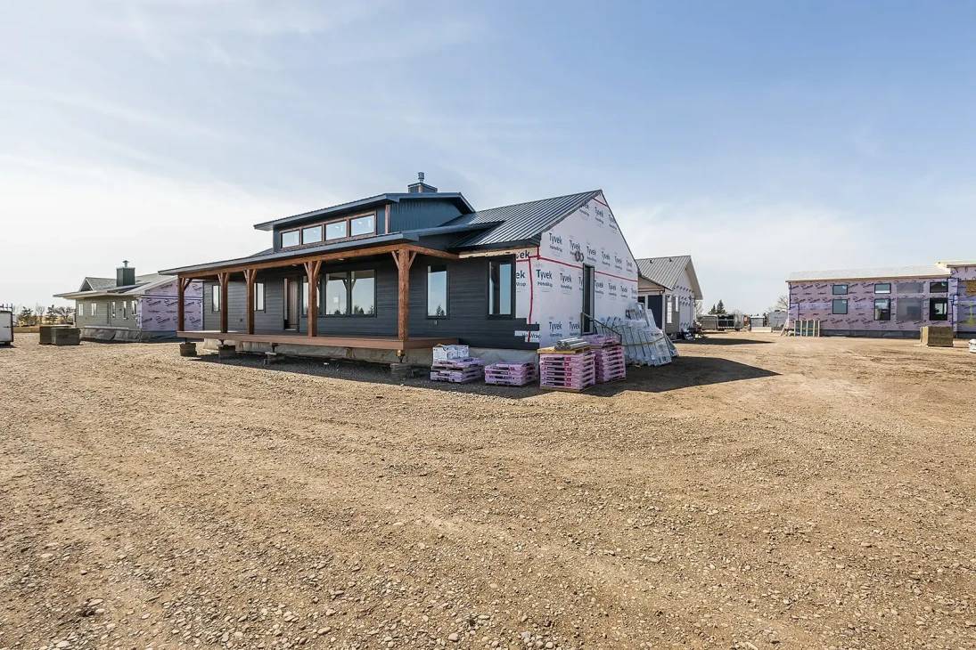 A partially constructed modern farmhouse with a dark roof sits on a dusty lot. Building materials are stacked nearby, under a clear blue sky.