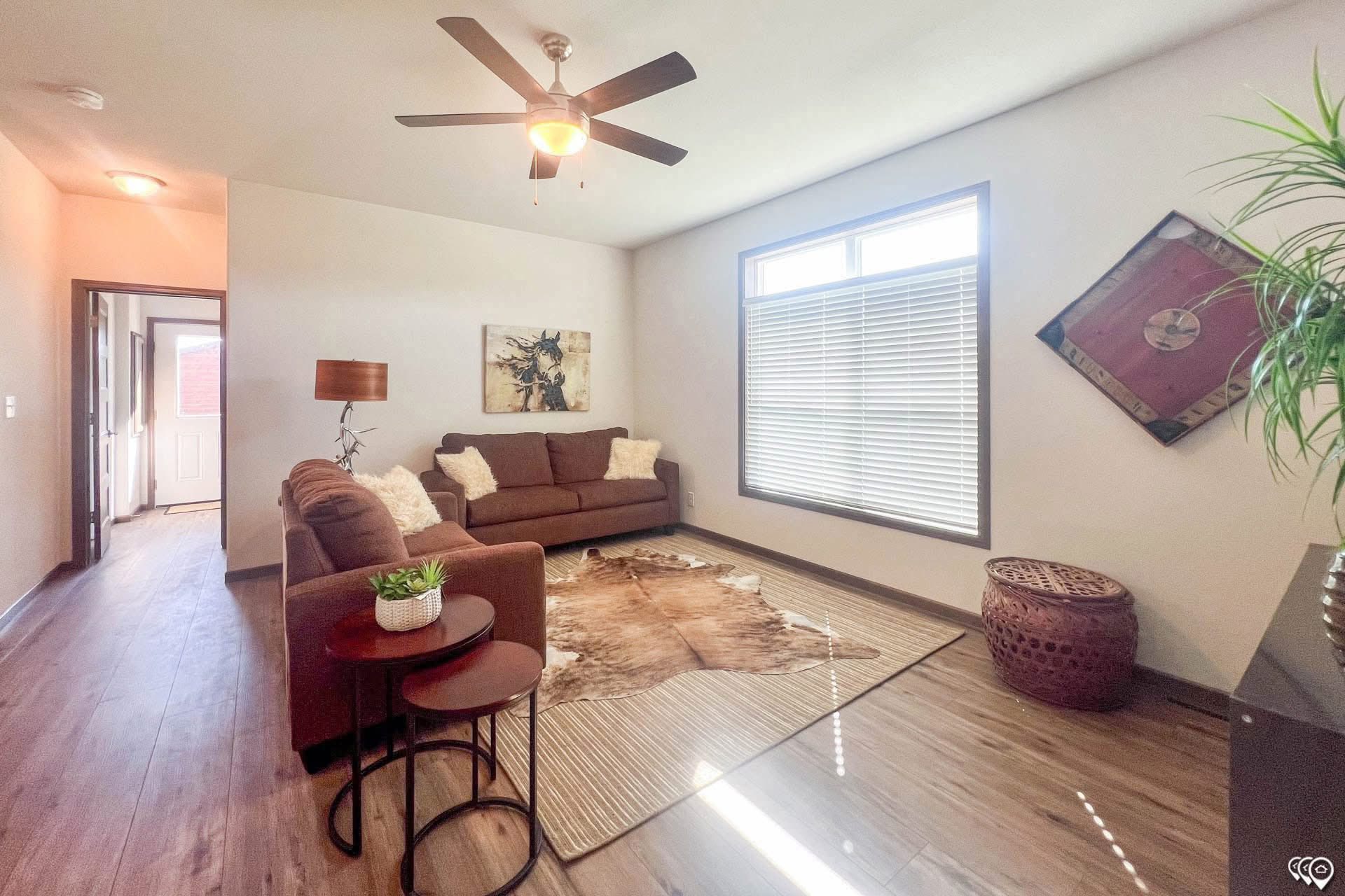 A cozy living room features a brown sofa with white pillows, a cowhide rug, and wooden floors. A ceiling fan spins above, and sunlight filters through a large window.
