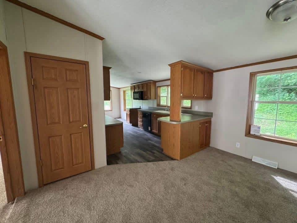 A cozy kitchen in a home with wooden cabinetry, dark countertops, and a carpeted area. Large windows let in natural light, revealing green scenery.