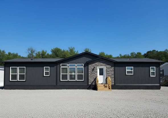 A modern, dark gray modular home with a stone-accented front, large windows, and a small wooden staircase. It's set against a bright blue sky and trees.