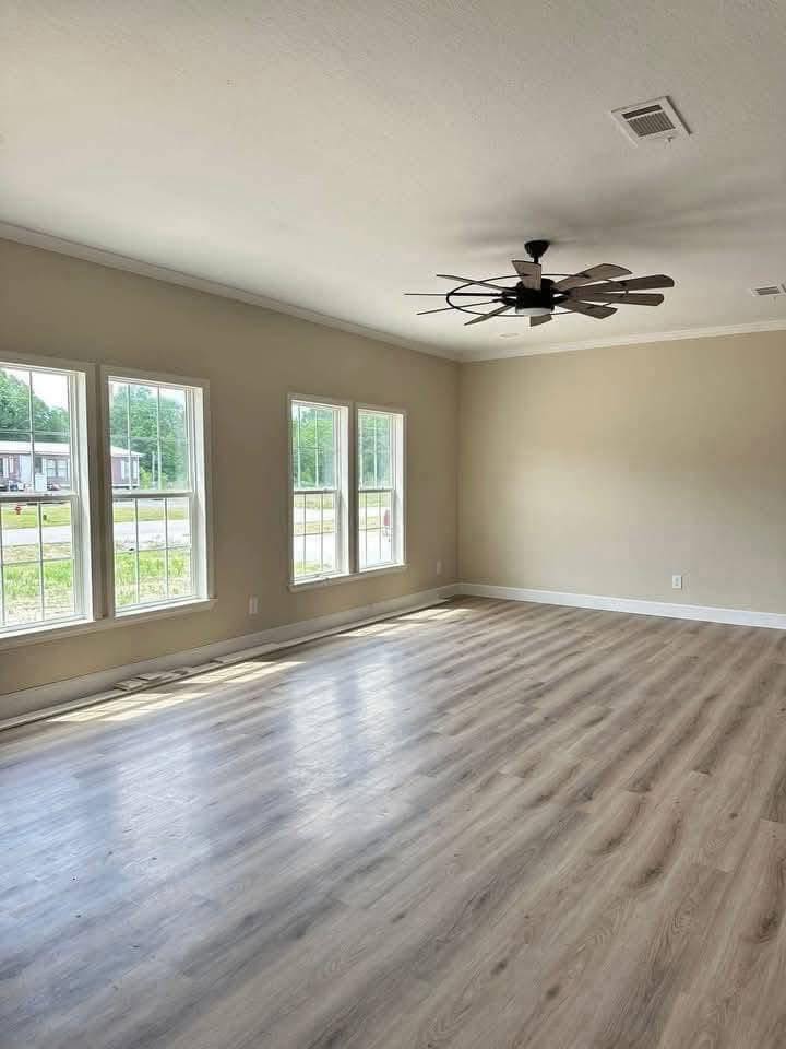 Spacious, empty room with light wood flooring, cream walls, and large windows letting in natural light. A modern ceiling fan hangs centrally.