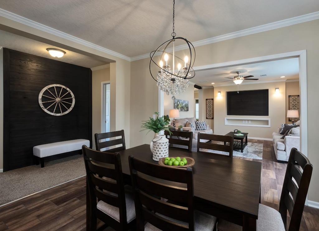 Elegant dining room with dark wooden table and chairs, a decorative chandelier above. Green apples and a vase adorn the table. Cozy living room in the background.