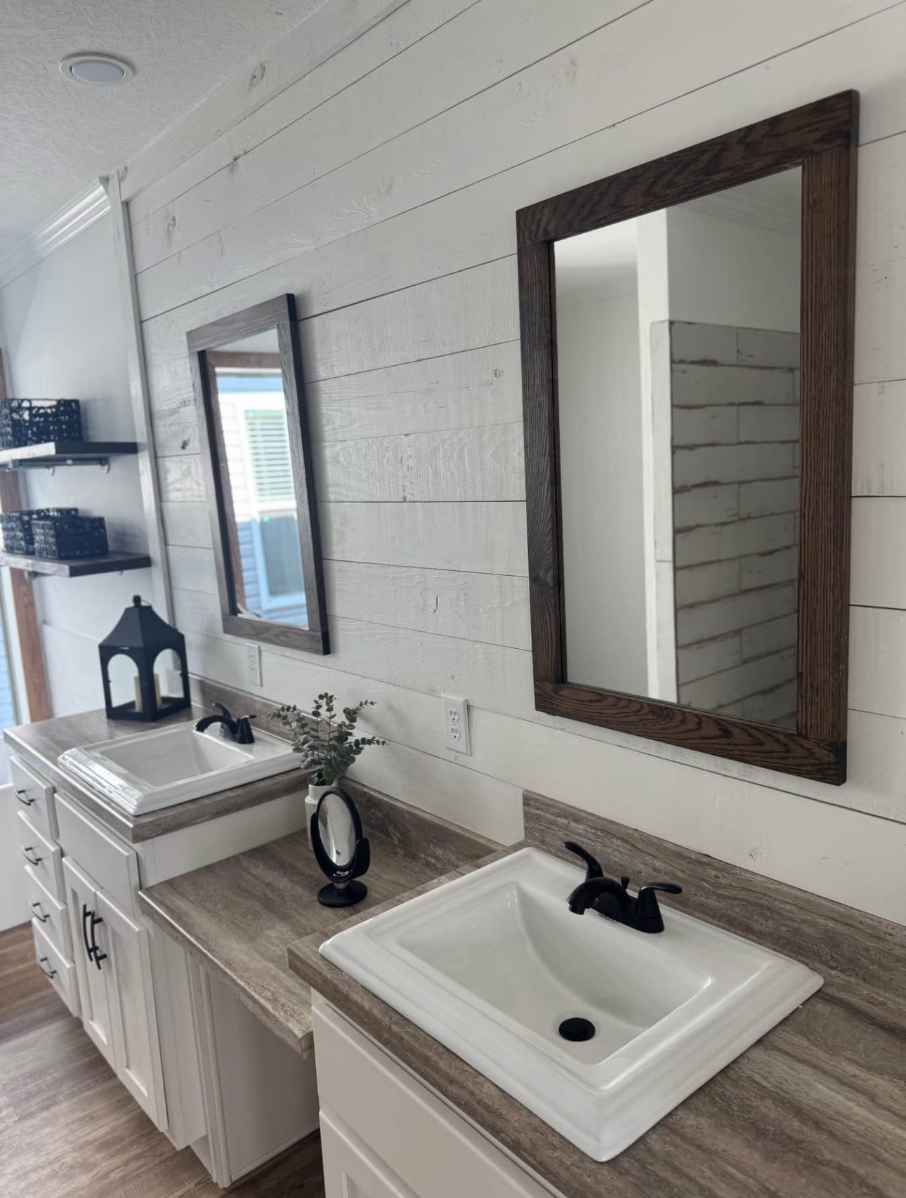 Modern farmhouse bathroom with two white sinks, dark faucets, and wood-framed mirrors on a shiplap wall. Shelves and decor add a rustic touch.