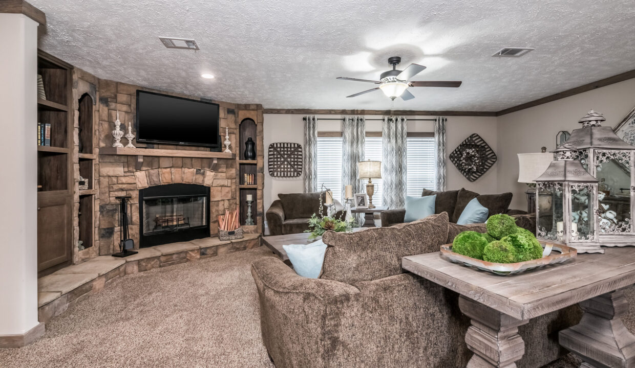 Cozy living room with brown sofas, stone fireplace, and a mounted TV. Soft lighting from a ceiling fan and table lamps adds warmth. Elegant decor throughout.