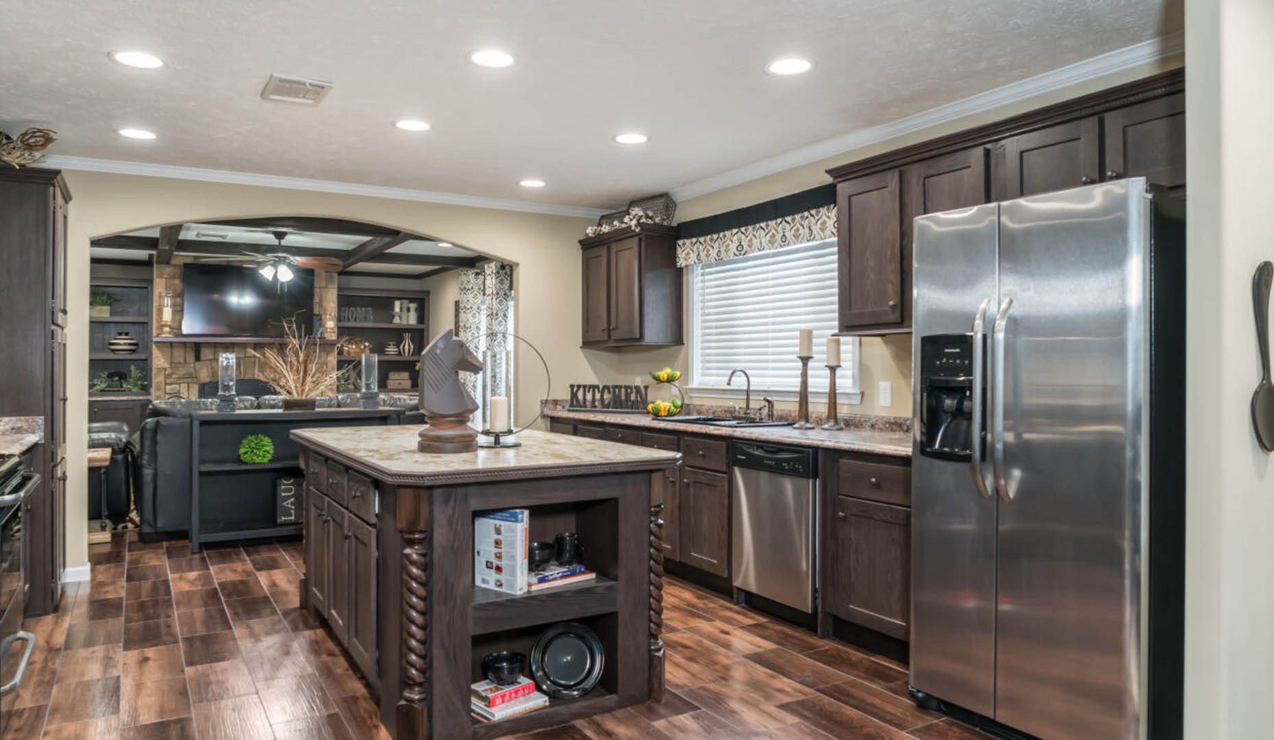 Modern kitchen with dark wood cabinetry and stainless steel appliances, including a refrigerator and dishwasher. Central island with decor, large window, and wood flooring create a warm, inviting atmosphere.
