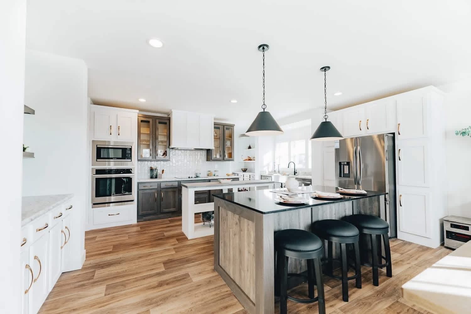 Modern kitchen with wooden flooring, white cabinets, black countertops, and stainless steel appliances. Three black stools at a central island create a cozy, inviting atmosphere. Bright lighting enhances the clean, spacious design.