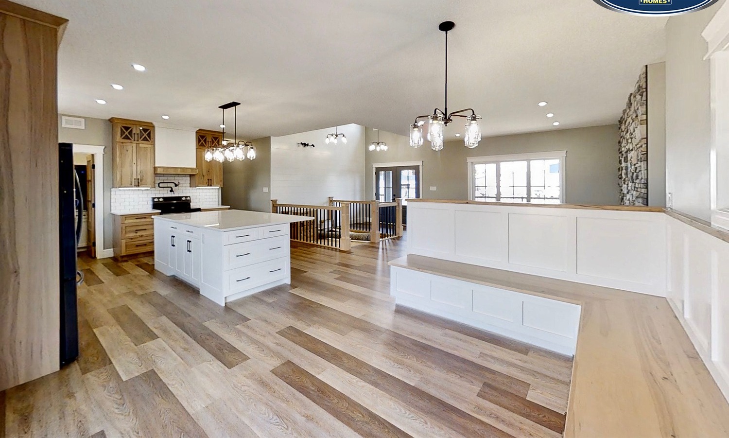 Spacious modern kitchen with light wood floors, white island, and pendant lights. Adjacent seating area with built-in bench and stone accent wall.