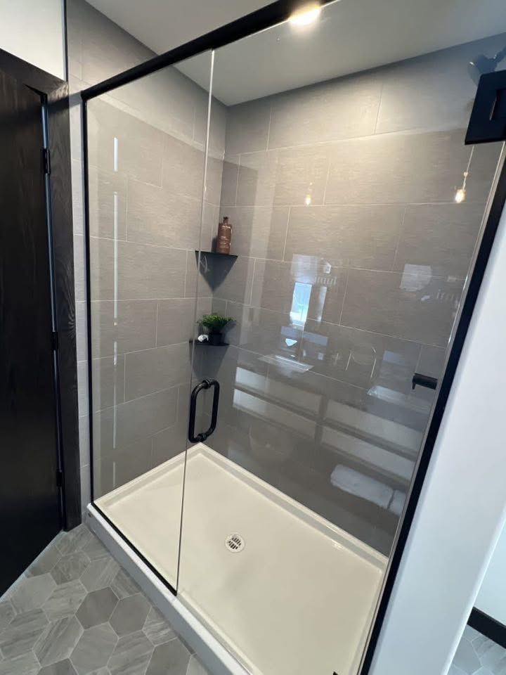 Modern bathroom with glass shower door, gray tiled walls, and hexagonal floor tiles. Inside, a corner shelf holds a small plant and a brown bottle.