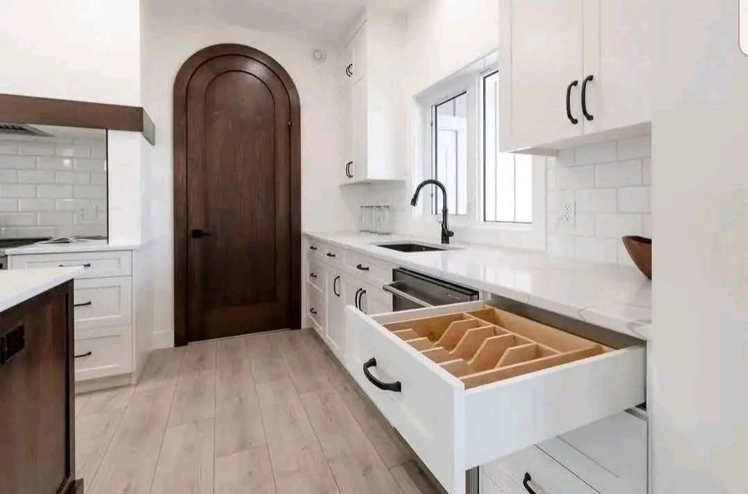 Sleek kitchen with white cabinets, a large arched wooden door, and a black faucet. A utensil drawer is open, revealing dividers. Bright and modern.