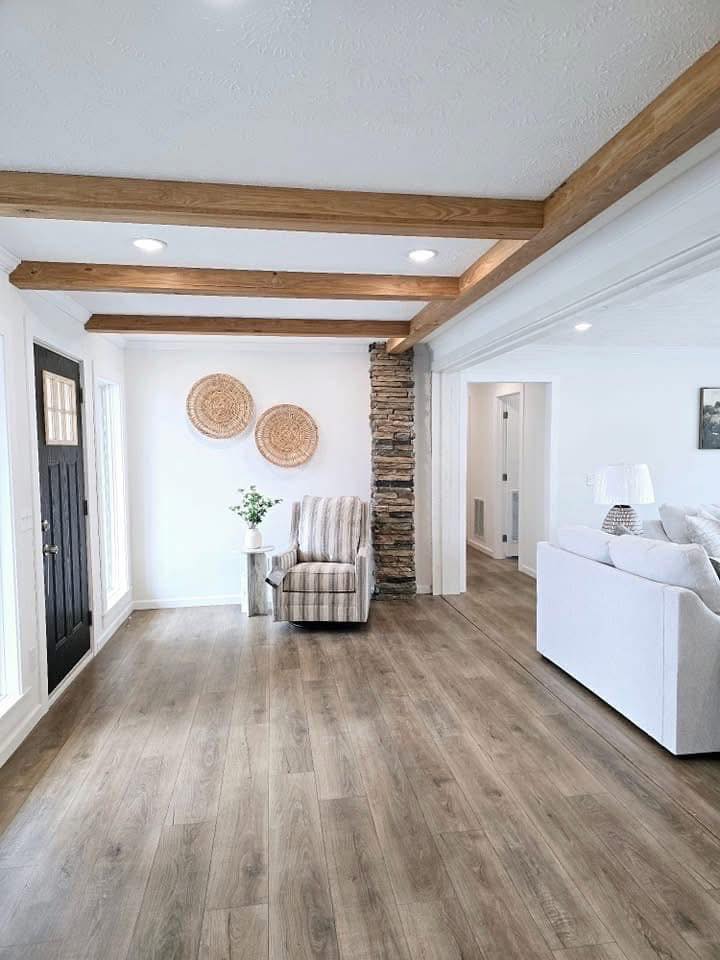 Bright living room with wood beams and stone column. Features a striped armchair, wicker wall decor, and a vase of flowers on a side table. Cozy ambiance.
