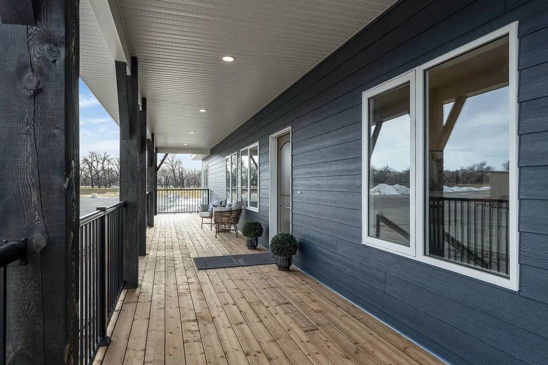 Covered porch with light blue siding, wooden floor, and black railing. Features a small bench and potted plants. Serene rural setting with trees.