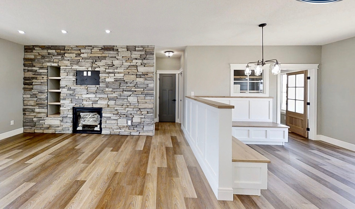 A modern living room with a stone accent wall featuring built-in shelves and a fireplace. Light wood flooring complements the neutral gray walls. A small dining area with a chandelier is adjacent, leading to a door with glass windows, creating an open, inviting atmosphere.