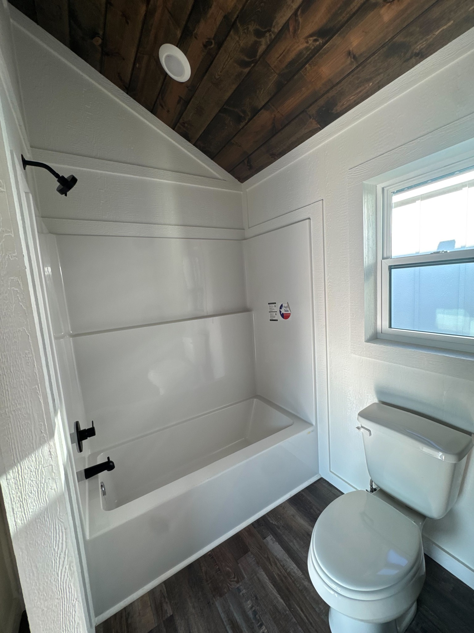 Compact bathroom with a white bathtub-shower combo, black fixtures, a toilet, and a small window. Wooden ceiling and vinyl plank flooring. Cozy and modern.