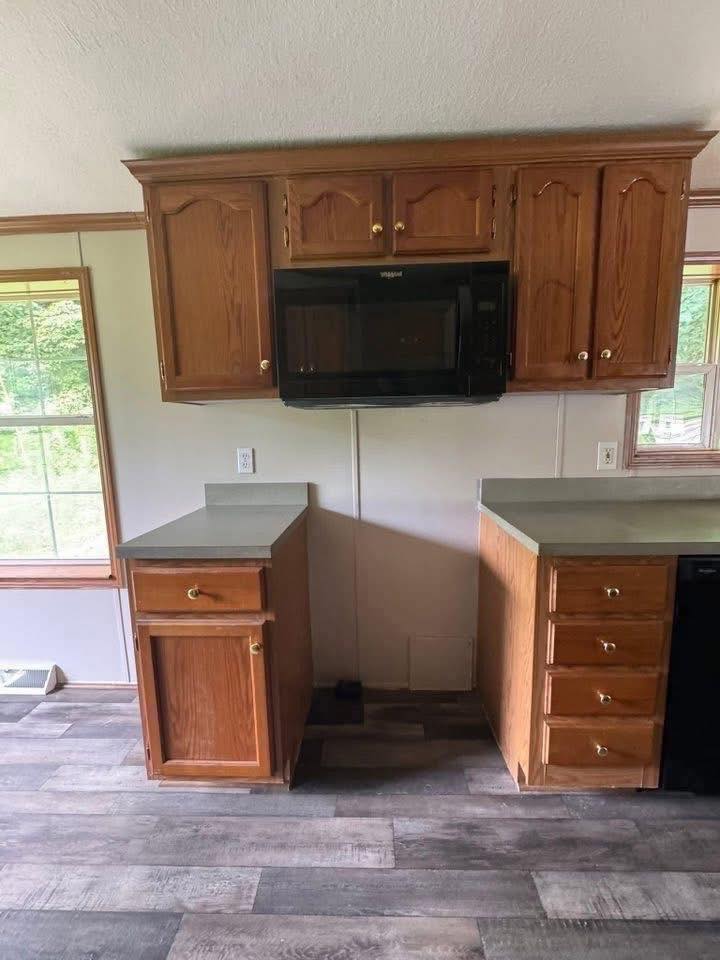 A kitchen area with wooden cabinets, a black microwave, and two small countertop sections. The room has wood-paneled flooring and large windows.