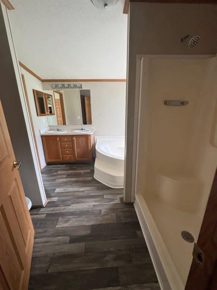 A bathroom with a wood floor, featuring a shower stall on the right, a double-sink vanity with mirrors on the left, and a large white bathtub.