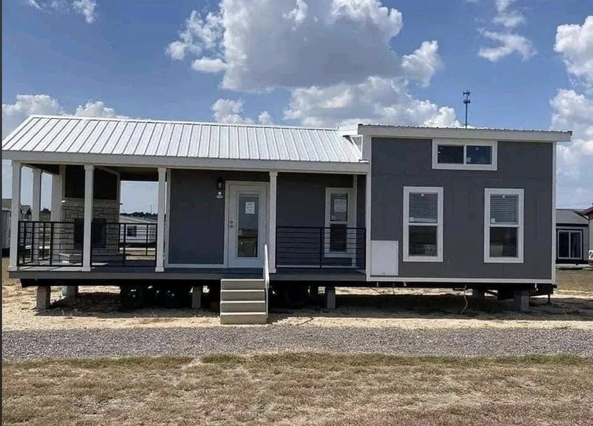 A modern, small gray house with white trim and a metal roof stands under a partly cloudy sky. It features a porch and steps leading to the entrance.
