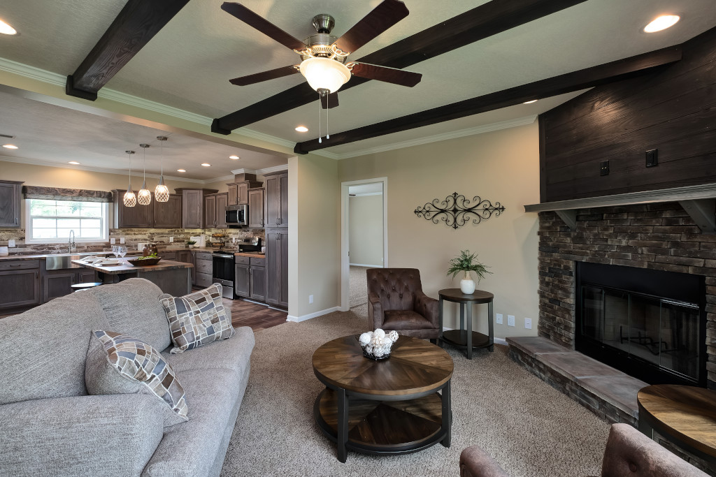 Cozy living room with beige walls, dark wood beams, and a ceiling fan. Features a gray sofa, stone fireplace, brown armchair, and open kitchen in the background.