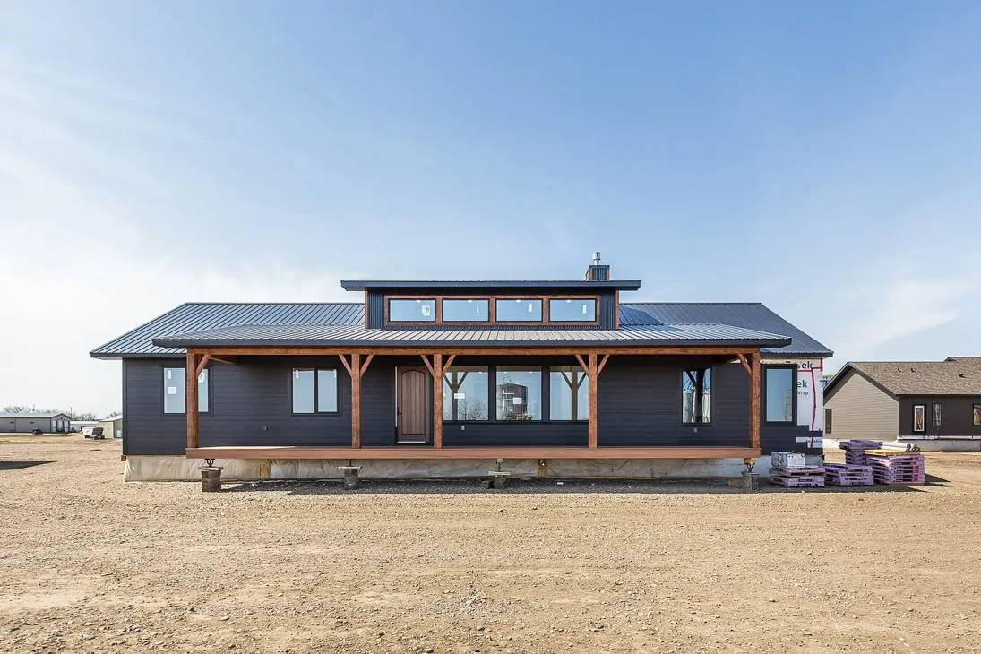 Single-story modern house with a dark facade, large windows, and wooden accents. Situated on barren land, under a clear blue sky, conveying simplicity.
