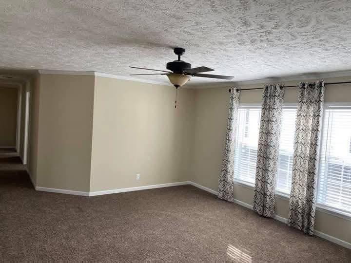 Spacious, empty living room with beige walls, carpeted floor, and two large windows covered by patterned curtains. A ceiling fan hangs from the textured ceiling. Cozy and inviting atmosphere.