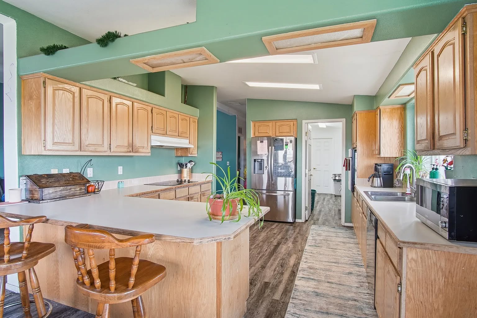 A spacious kitchen with green walls features light wooden cabinets and white countertops. Two wooden stools sit at the counter, while potted plants and appliances add a cozy touch. The room is bright and welcoming.