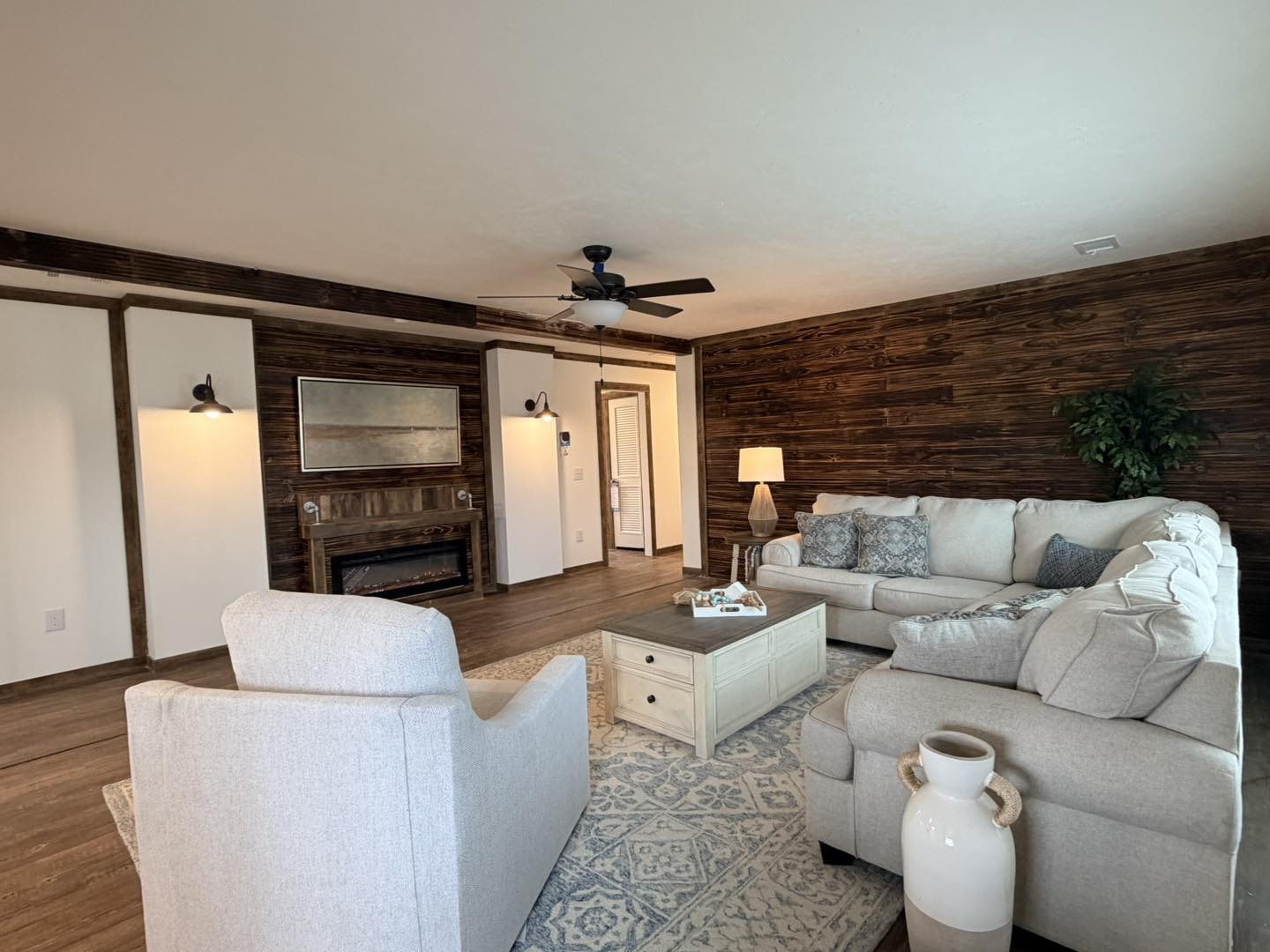 Cozy living room with a beige sectional sofa and armchair, wooden coffee table, and rustic wooden walls. Warm lighting and ceiling fan convey comfort.