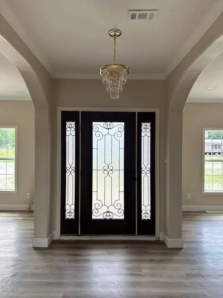 Elegant entryway with a dark wood door featuring ornate glass panels, flanked by bright windows. A chandelier hangs from the ceiling, casting a refined ambiance.