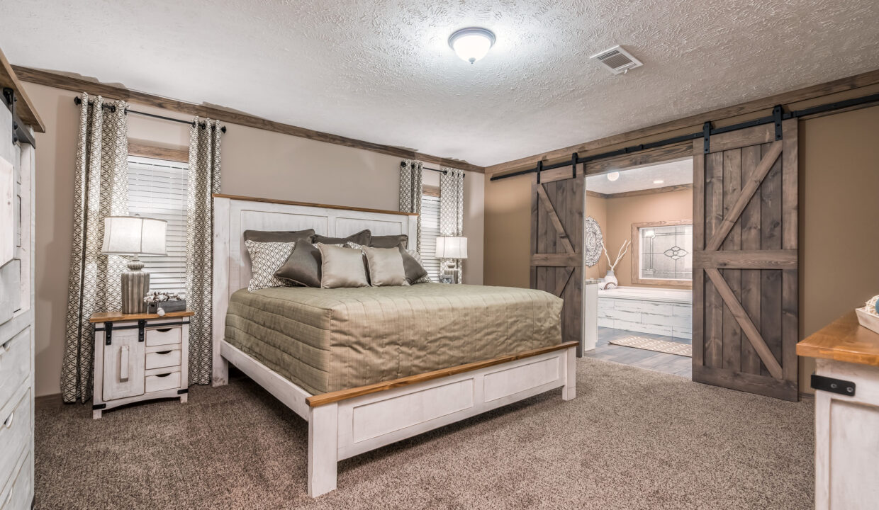 Cozy bedroom with a king-sized bed, wooden headboard, and green bedding. Soft lighting, earth tones, barn doors lead to a bath, creating a rustic feel.