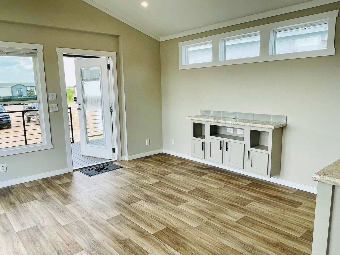 Bright living room with light wood flooring, beige walls, and high windows. Features a built-in cabinet and open glass door leading outside. Modern and airy.
