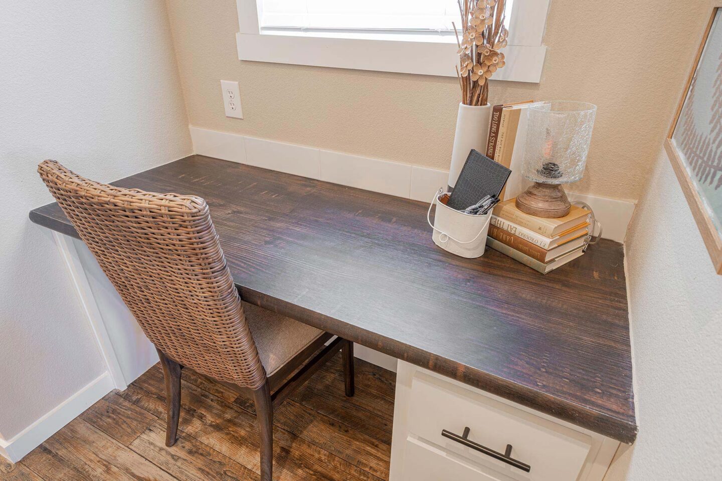 A cozy workspace features a wicker chair at a dark wooden desk with books, a lamp, and decorative items. A window above lets in soft light, lending a serene feel.