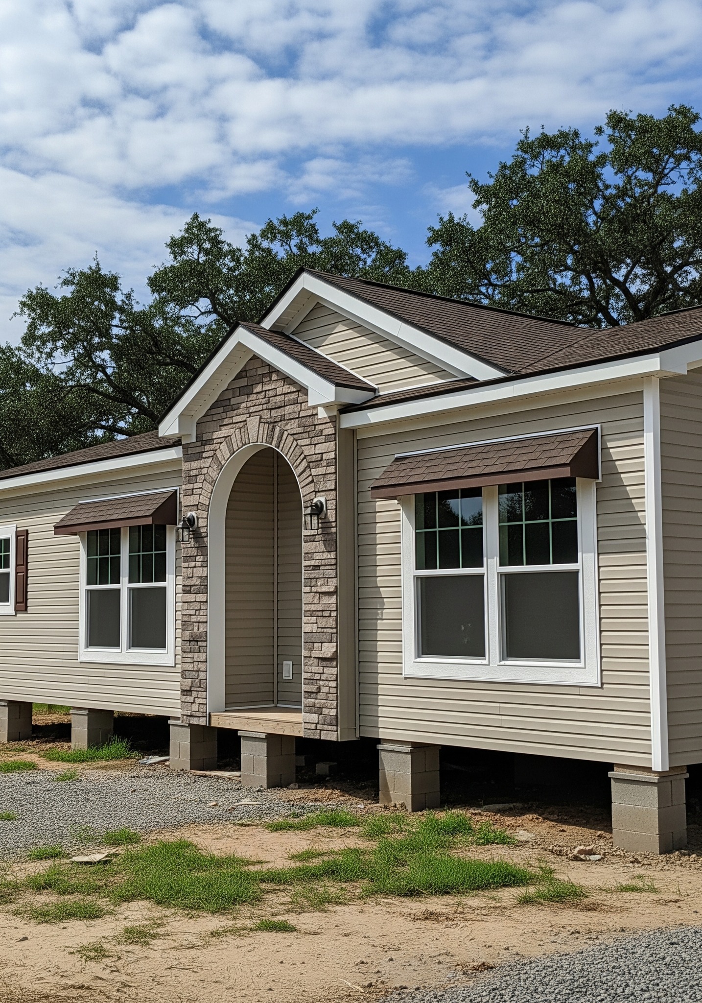 A beige manufactured home on blocks, featuring a stone archway, white-trimmed windows, and brown awnings. Set against a backdrop of green trees and a cloudy sky.