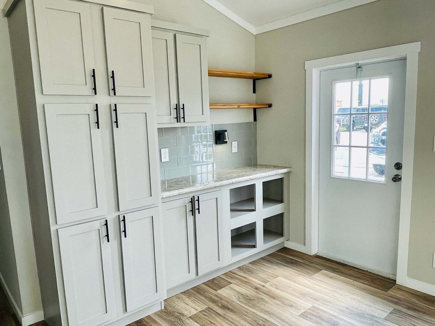 Modern kitchen with light gray cabinets, black handles, open wooden shelves, stone countertop, and glass door. Light wood floor adds warmth.