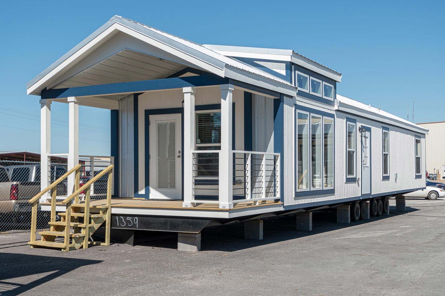 Modern mobile home with blue and white exterior, large windows, and a small porch with steps. The home is parked on a paved lot under a clear blue sky.